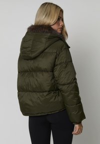 Kobieta z blond w�łosami w oliwkowej kurtce puchowej z futrzanym kapturem, stojąca bokiem na jednolitym tle.