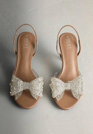 Beige Sandalen mit offener Zehenpartie, klarem Riemen und großer, strukturierter Schleife, die mit klaren Perlen und Pailletten verziert ist, auf einem glatten Hintergrund liegend.