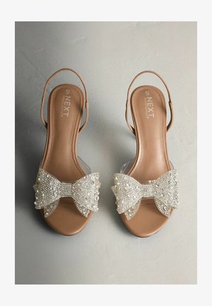 Beige Sandalen mit offener Zehenpartie, klarem Riemen und großer, strukturierter Schleife, die mit klaren Perlen und Pailletten verziert ist, auf einem glatten Hintergrund liegend.