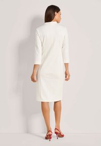 Weißes, tailliertes Kleid mit Dreiviertelarmen, hohem Ausschnitt und einer Mittelnaht hinten. Getragen mit roten High Heels. Glatte Textur.