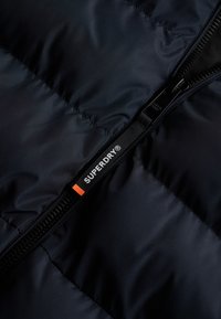 Donkerblauwe pufferjack met een gladde textuur en een zichtbaar gestikt patroon. Voorzien van een zwarte rits en een logo-label met oranje accenten.