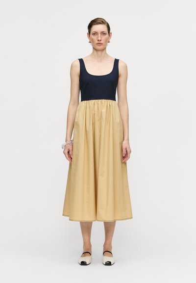 Marineblaue ärmellose Kleid mit einem beigen, knielangen Rock. Glattes Material, lässige Passform und geraffte Taille. Schwarz-weiße Slipper.