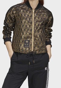 Černá bomber bunda s geometrickým zlatým vzorem, vybavená předním zipem, žebrovanými manžetami a štítkem s logem značky na dolní části předního dílu.