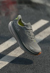 Scarpa da corsa HOKA leggera in mesh grigio, con accenti giallo neon, suola ammortizzata e dettagli riflettenti per una maggiore visibilità.