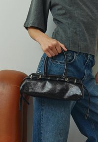 Geantă de piele neagră cu un finisaj texturat, mânere scurte și o formă compactă, ținută împotriva unui top gri și blugi bleu.