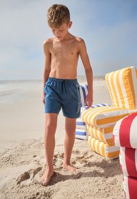 Pantaloncini da bagno blu con coulisse, caratterizzati da un logo sul lato. A piedi nudi sulla sabbia della spiaggia con sedie a strisce colorate sullo sfondo.