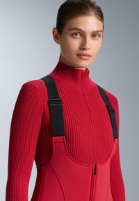 Piros bordázott pulóver magas gallérral, piros overállal párosítva, amely fekete állítható pántokkal és cipzáras záródással rendelkezik. Simább tapintású anyag.