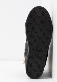 Baskets noires avec une tige texturée et des ferrures dorées. Elles présentent une semelle en caoutchouc à motif, étiquetée avec "GUESS", inclinée sur un fond blanc.