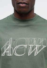 Tricou din bumbac de culoare verde măslin, cu litere mari, alb trasate cu linia, "4CW", afișate proeminent pe față. Design clasic cu guler rotund.