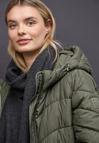 Chaqueta de plumas verde oliva con capucha, que presenta un acabado texturizado y un patrón cosido, llevada sobre una bufanda gris oscuro.