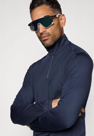 Camisola de manga longa azul-marinho com fecho éclair e colarinho alto, feita de um tecido liso e ajustado. O modelo usa óculos de sol reflectores oversized com uma forma geométrica.