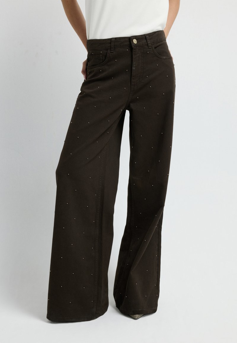 Pantaloni a gamba larga marroni con vita alta, decorati con piccoli chiodi metallici, realizzati in morbido cotone, con il classico design a cinque tasche.