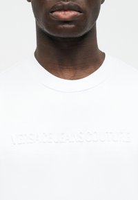 Κοντινό πλάνο ενός ατόμου που φορά λευκό μπλουζάκι με στρογγυλή λαιμόκοψη και ανάγλυφο κείμενο "Versace Jeans Couture" στο στήθος.