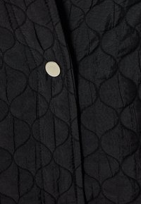 Tessuto di cotone nero trapuntato con un motivo a onde, fissato con un bottone circolare argento, che enfatizza la texture e i dettagli del design.
