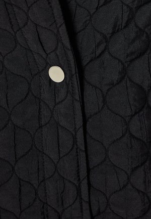 Sort quiltet stof med et bølget mønster, fastgjort med en cirkulær sølvknap, der fremhæver tekstur og design detaljer.