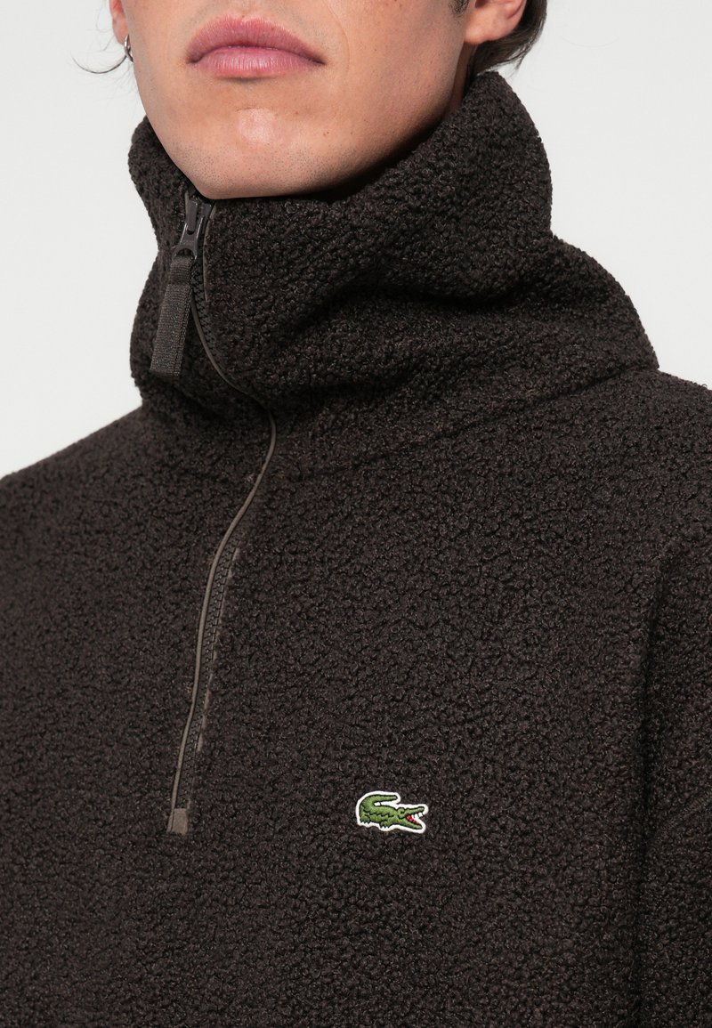 Fleece-pullover i mørkebrun, med høj krave, kvart-zip lukning og et grønt broderet krokodillelogo på brystet.