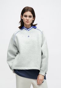 Νεαρός με κοντά καστανά μαλλιά που φοράει ένα γκρι φούτερ πάνω από ένα μπλε πουκάμισο και ανοιχτόχρωμα παντελόνια, ποζάροντας μπροστά από απλό φόντο.