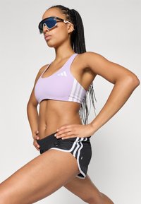 Sostén deportivo de lavanda claro con tres rayas blancas y un logo; combinado con pantalones cortos deportivos negros con ribetes blancos y un ajuste relajado.