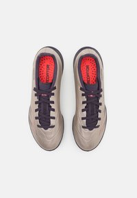 Chaussures de sport beige et violette avec une tige texturée, semelle noire et accents rouges à l'intérieur. Équipées de lacets classiques et d'une marque.