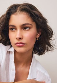 Gold heart-shaped earrings are worn, complemented by a white linen shirt. The model has medium-length, wavy dark hair and natural makeup.