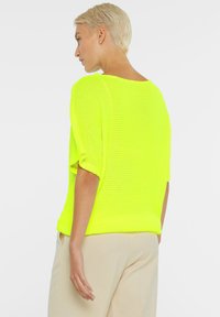 Neon-gelbes gestricktes Sweatshirt mit kurzen Ärmeln und lockerer Passform, mit rundem Halsausschnitt und geripptem Saum, kombiniert mit beigen Hosen.