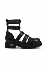 Botas de tobillo de cuero negro que presentan lados recortados, múltiples correas, detalles perforados, un tacón grueso y una cremallera lateral para facilitar el uso.