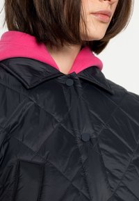 Schwarze gesteppte Jacke mit einer rosa Fleecekapuze, die einen spitzen Kragen und zwei schwarze Knöpfe vorne hat. Glatte, glänzende Textur.