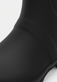 Μαύρη ραφινάτη μπότα μέχρι τον αστράγαλο από ελαστικό υλικό με ματ φινίρισμα, στρογγυλή μύτη και εύκαμπτο επάνω μέρος με ελάχιστο σχεδιασμό και χωρίς εμφανή μεταλλικά στοιχεία.