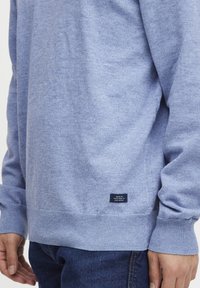 Sweatshirt albastru deschis, realizat dintr-un material moale, cu mansete și tiv cu nervuri, având un eticheta mică pe partea stângă jos.