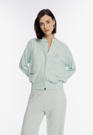 Kobieta z krótkimi ciemnymi włosami, ubrana w jasnobłękitną kurtkę bomberkę i dopasowane spodnie, patrząca prosto, ręce z tyłu, na białym tle.