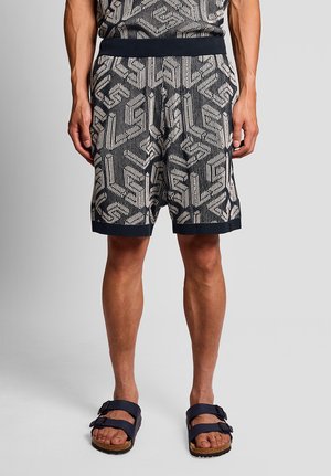 Man draagt marineblauwe gebreide korte broek met patroon en trekkoord, en zwarte sandalen met gespen, op een grijze achtergrond.