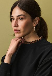 Tricou negru cu guler de leopard texturat și cu volane. Modelul are părul brunet, cercei aurii și un fundal neutru.