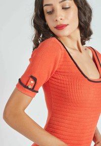 Vestido rojo de canalé con mangas cortas, escote cuadrado, ribete negro y detalles de botones dorados en las mangas. Diseño ajustado con tejido texturizado.