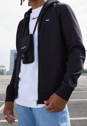 Person trägt eine schwarze Zip-up-Hoodie, weißes T-Shirt, hellblaue Jeans und eine schwarze Umhängetasche mit dem "Tommy Jeans"-Logo im Freien.