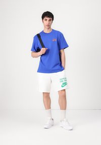 Mėlyna trumpa rankovė marškinė su oranžiniu logotipu, suderinta su baltais šortais, kuriuose yra žalias tekstas ir ženklai, papildyta baltais sportbačiais.
