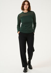 Groene, sprankelende blouse met lange mouwen en een ronde hals, gecombineerd met zwarte, wijde broek en zwarte schoenen, wat een slanke silhouet creëert.