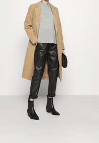 Osoba w beżowym płaszczu, szarym swetrze, czarnych skórzanych spodniach, czarnych botkach za kostkę, trzymająca czarną kopertówkę, stojąca na białym tle.