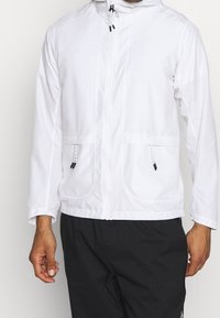 Chaqueta blanca con capucha que presenta un cierre frontal, dos bolsillos delanteros, cintas ajustables y puños elásticos para un ajuste seguro.