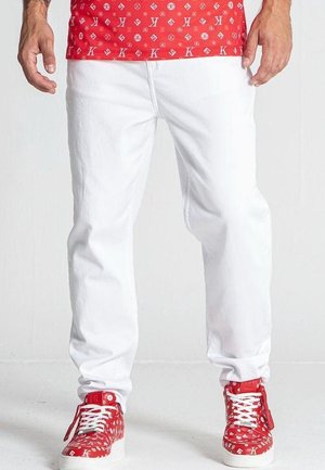 Mężczyzna w białych spodniach, czerwonych wysokich trampkach i czerwonej koszulce z białymi wzorzystymi literami i symbolami.