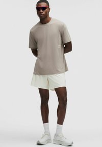 Mężczyzna stoi z rękami za plecami, ubrany w beżowy T-shirt, białe szorty, białe sneakersy, skarpety do kostek i sportowe okulary przeciwsłoneczne w czerwonym odcieniu na tle jednolitego tła.