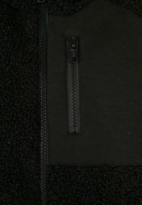 Zwarte fleece jacket met een glad zwart stoffen paneel, voorzien van een ritssluiting aan de voorkant en een ritszak op de borst.