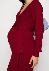 Femme enceinte portant une robe enveloppante à manches longues de couleur rouge foncé avec une ceinture nouée, portant un sac à main matelassé bleu clair sur son épaule.