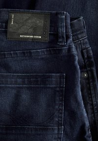 Tumed denimist valmistatud teksad, millel on tekstuuriga pind, must nahklabel, millel on kiri "RETHINKING DENIM" ning nähtavad õmblused.