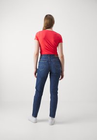 Červený krátky rukáv top v kombinácii s modrými džínsami. Džínsové nohavice majú klasický strih, viditeľné švy a dizajn zadnej vrecka. Biele tenisky dopĺňajú celkový vzhľad.