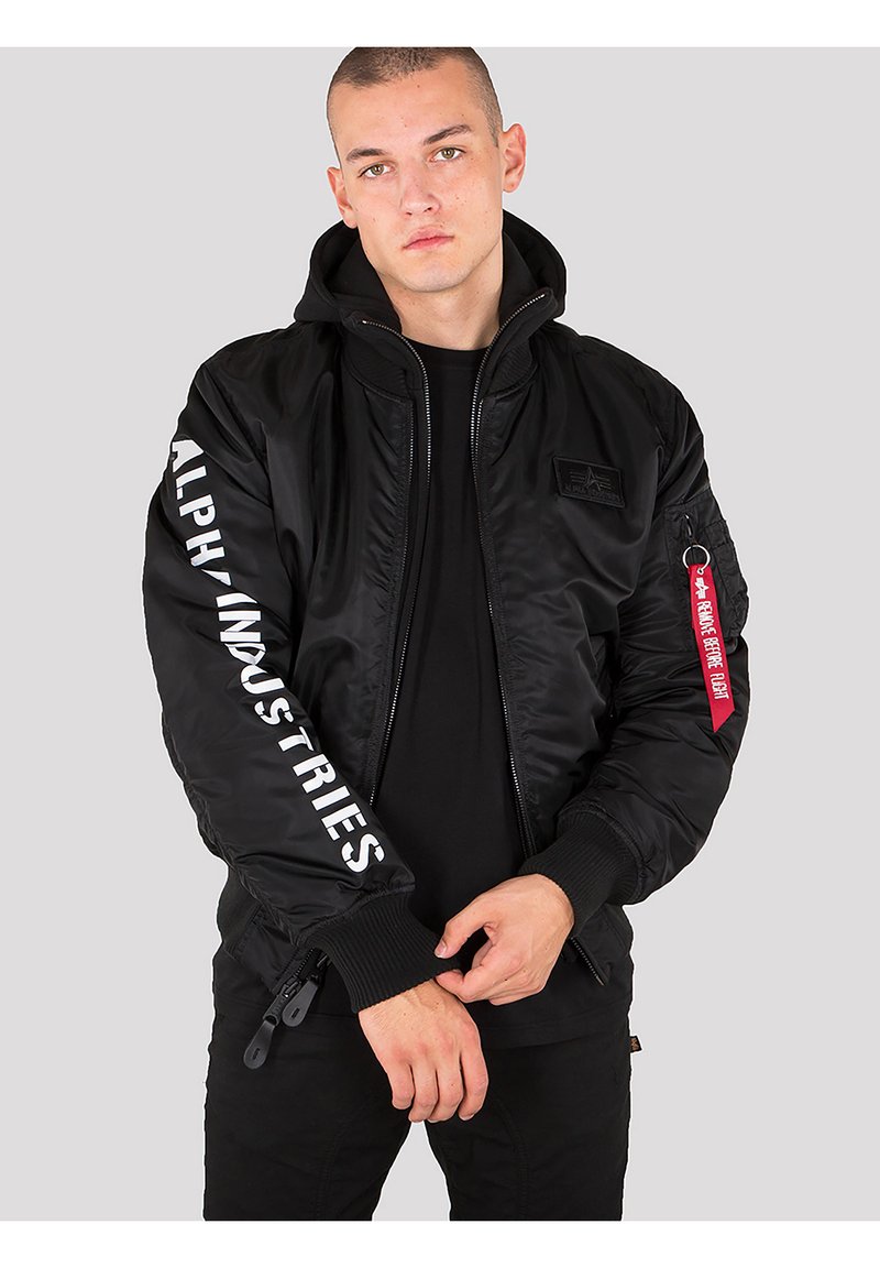 Čierna bombrová bunda s ribovanými manžetami, zipsom vpredu a s potlačou "ALPHA INDUSTRIES" v bielej farbe na rukáve. Obsahuje červenú látkovú etiketu na vrecku.