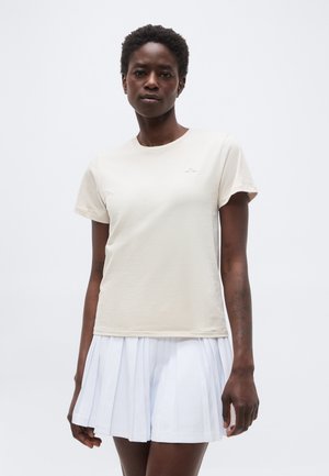 Γυναίκα με κοντά σγουρά μαλλιά, που φοράει ένα ανοιχτό μπεζ μπλουζάκι και λευκή πλισέ φούστα, στέκεται μπροστά σε απλό λευκό φόντο.