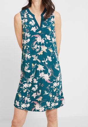 Kobieta w bezrękawniku zielonkawoniebieskiej sukience z różowym, białym i żółtym kwiatowym wzorem oraz dekoltem w kształcie litery V, stojąca na jednolitym tle.