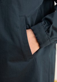 Ciemnoniebieska kurtka z boczną kieszenią i elastycznymi ribanami przy mankietach. Materiał wygląda na gładki o matowej teksturze. Projekt jest elegancki i minimalistyczny.