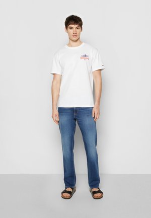 Hvid bomulds t-shirt med trykt "TOMMY'S" logo i rød og blå. Bæres med blå jeans og sorte sandaler. Enkel skæring og klassisk pasform.