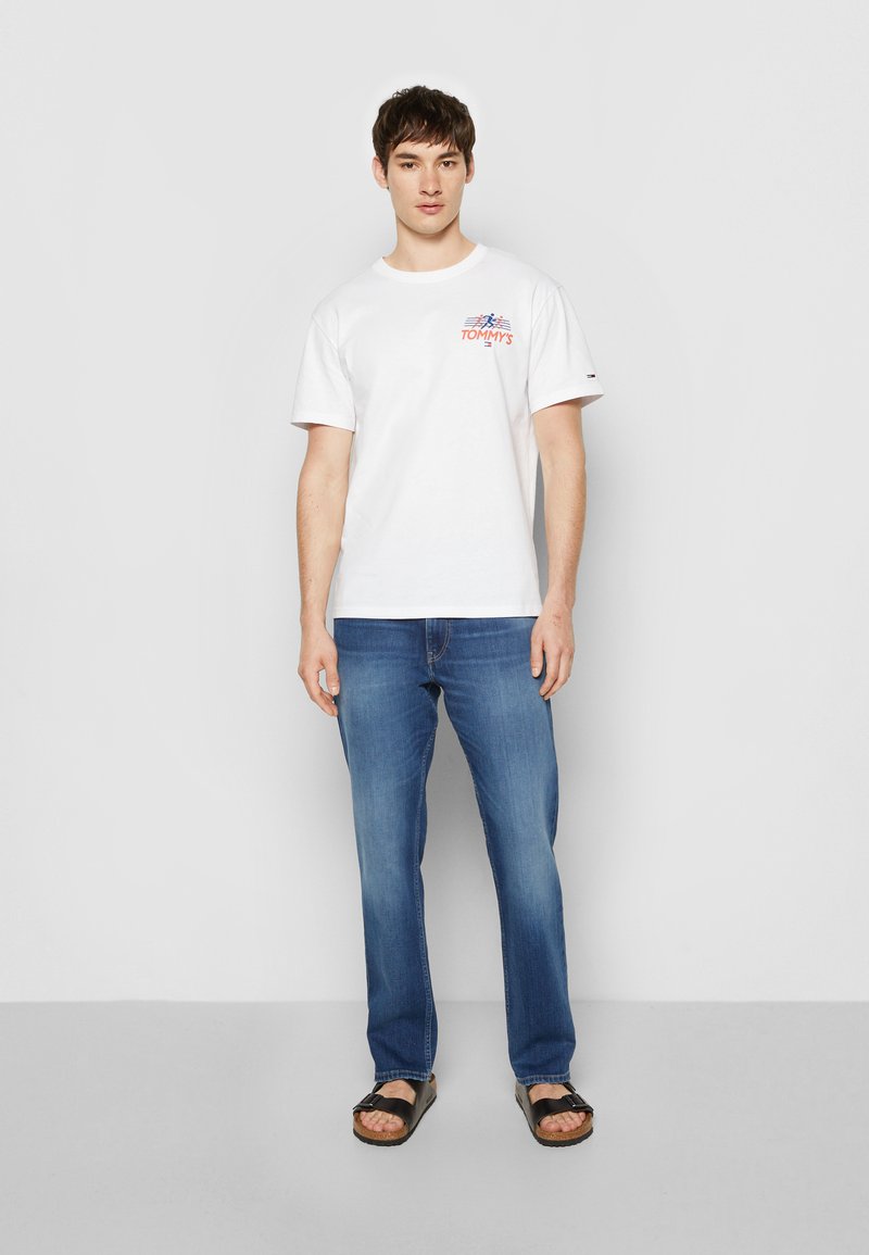 Hvid bomulds t-shirt med trykt "TOMMY'S" logo i rød og blå. Bæres med blå jeans og sorte sandaler. Enkel skæring og klassisk pasform.